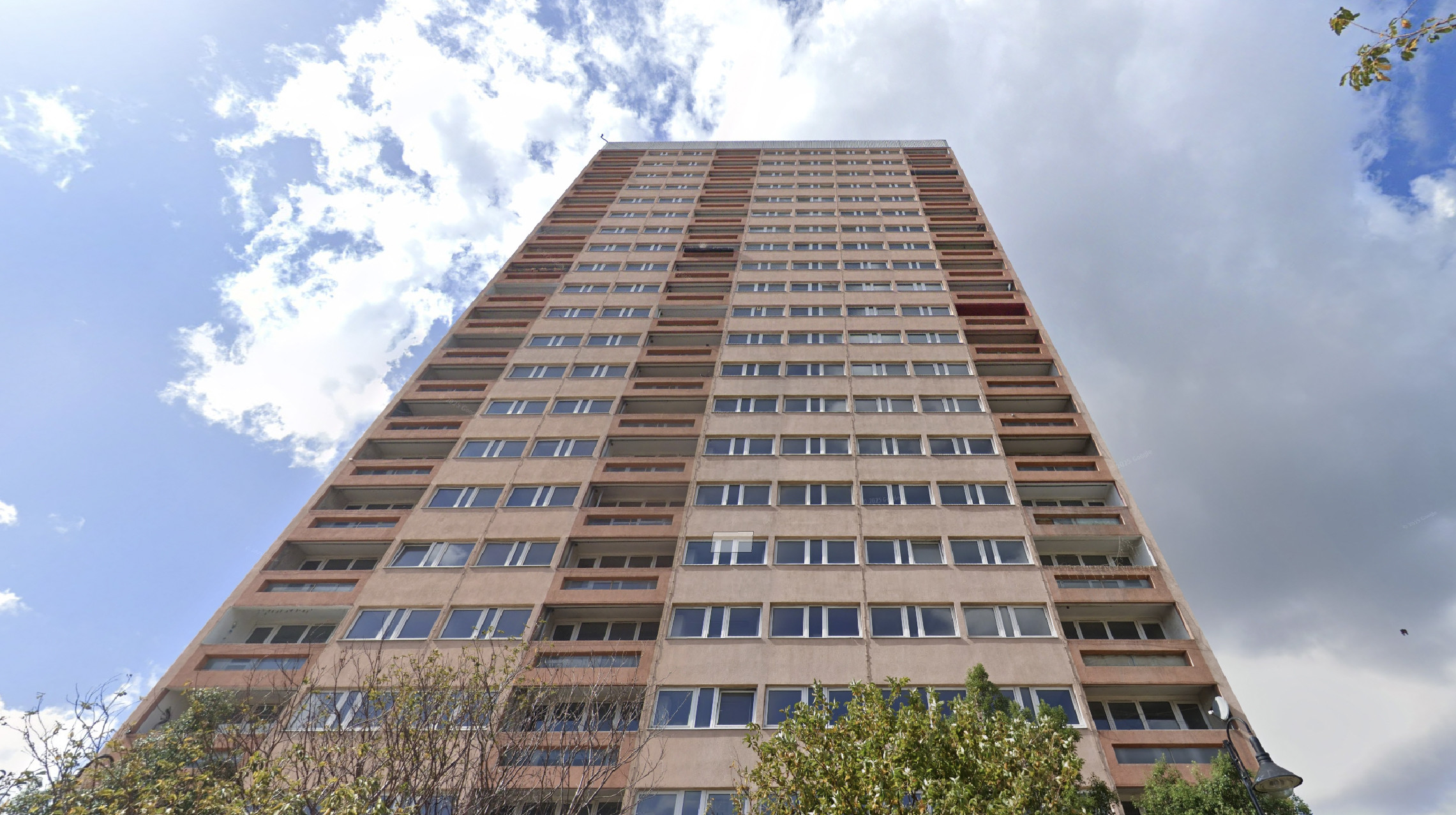 Low-angle view of the existing Clare House, a tall beige high-rise apartment building with multiple rows of windows and balconies under a partly cloudy sky, with trees visible at the base.